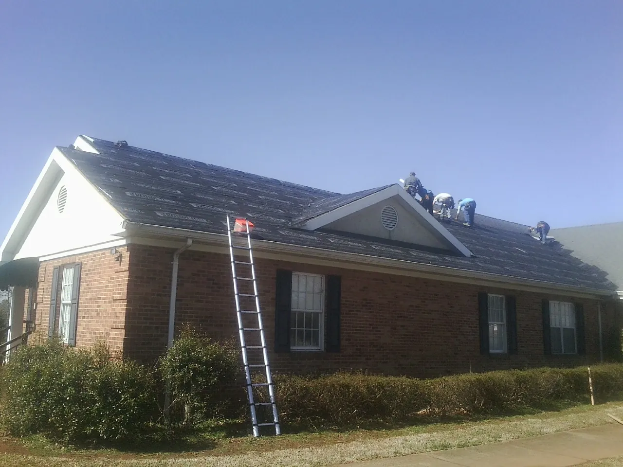 Crew tearing shingles off a brick ranch roof with a ladder set — Emergency Roof Repair work by our crew in Good Thunder