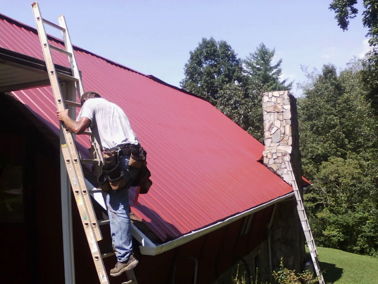 Roofer on a ladder beside a red metal roof and stone chimney — Soffit & Fascia Repair by Roofing Brothers in Good Thunder